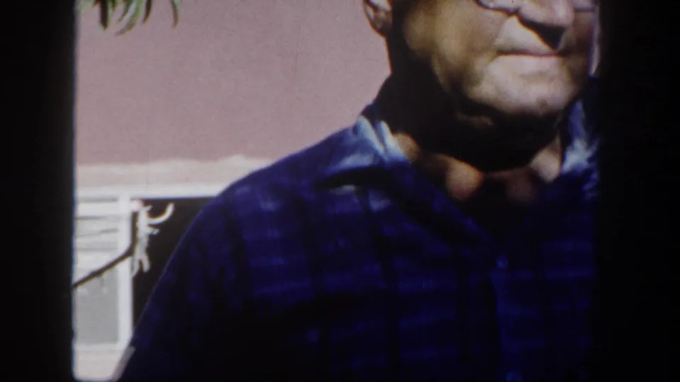 A closer, partly cropped view shows the older man in a dark checked shirt near a pink exterior wall and window in the supplied 1962 San Diego footage.