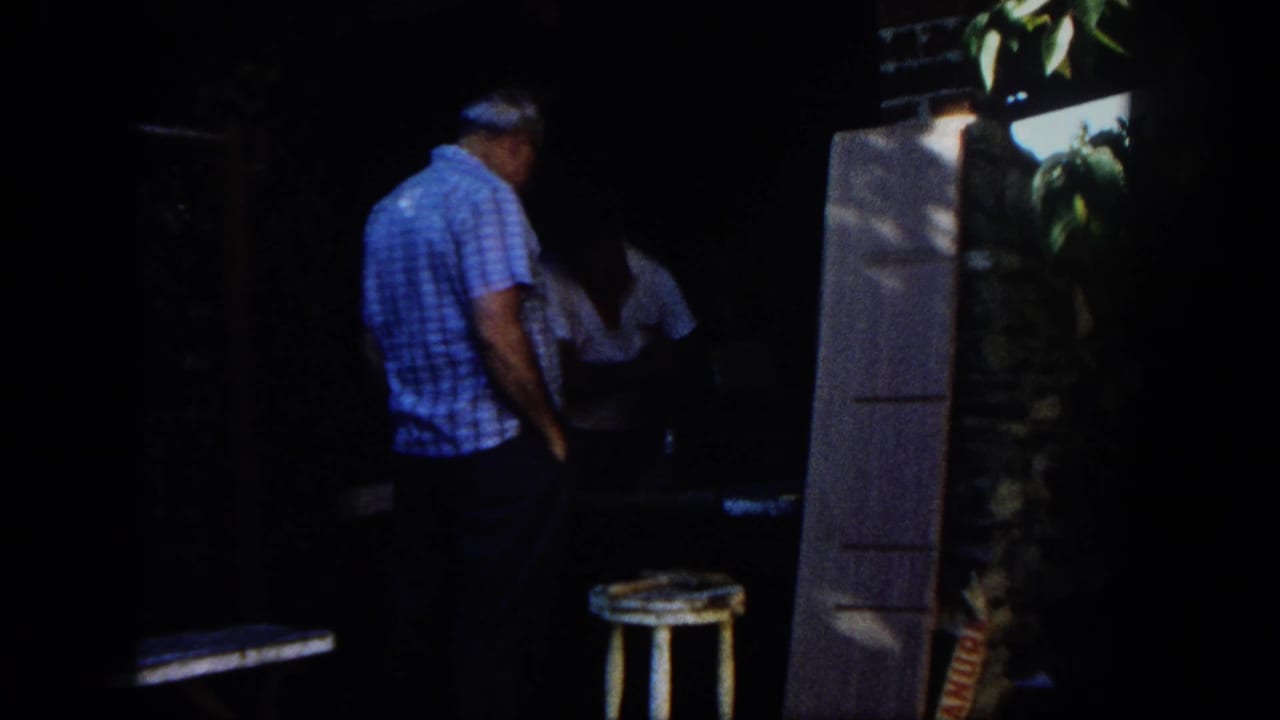 Two adult men in a shaded outdoor workshop, one standing near a table and stool.