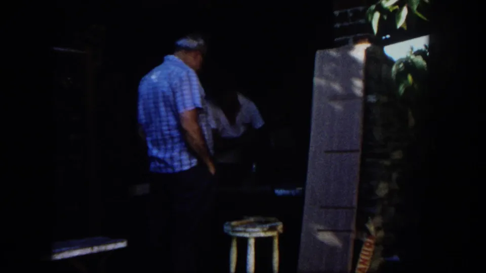 1962 San Diego home-movie frame of an adult man in a blue checkered shirt watching another man work at a table in a shaded outdoor workshop.