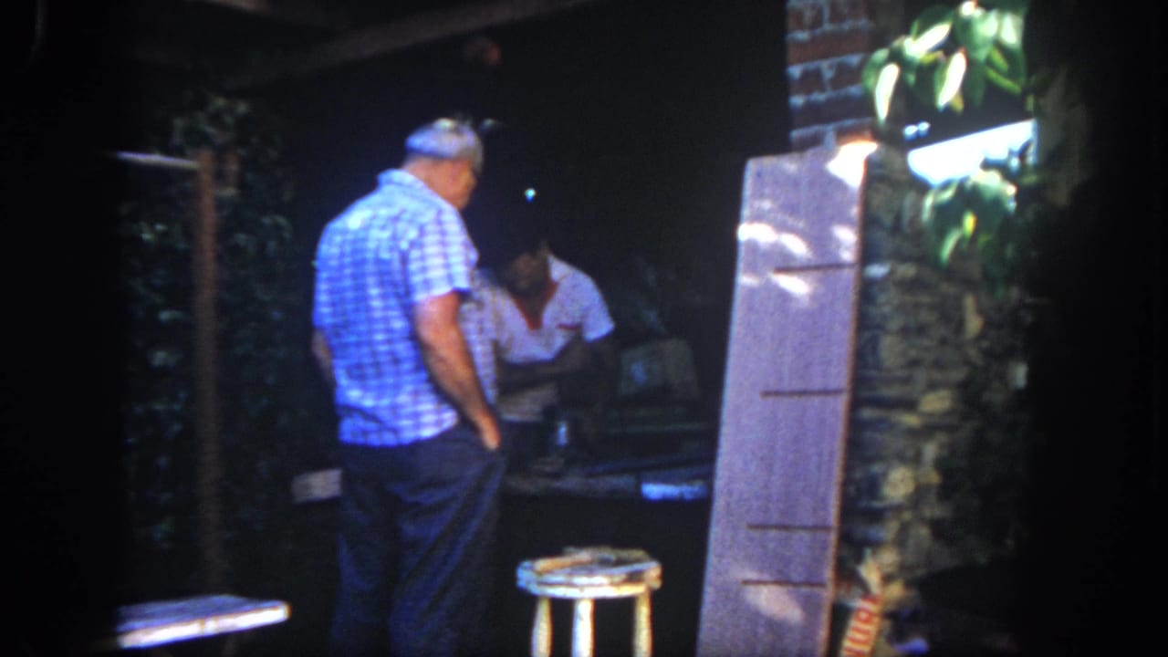 Man in a blue checkered shirt stands near a seated or leaning man at a worktable.