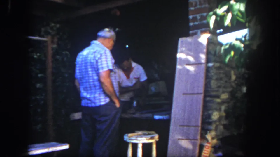 1962 San Diego outdoor workshop view with a man in a blue checkered shirt watching another adult at a dim worktable near boards and foliage.