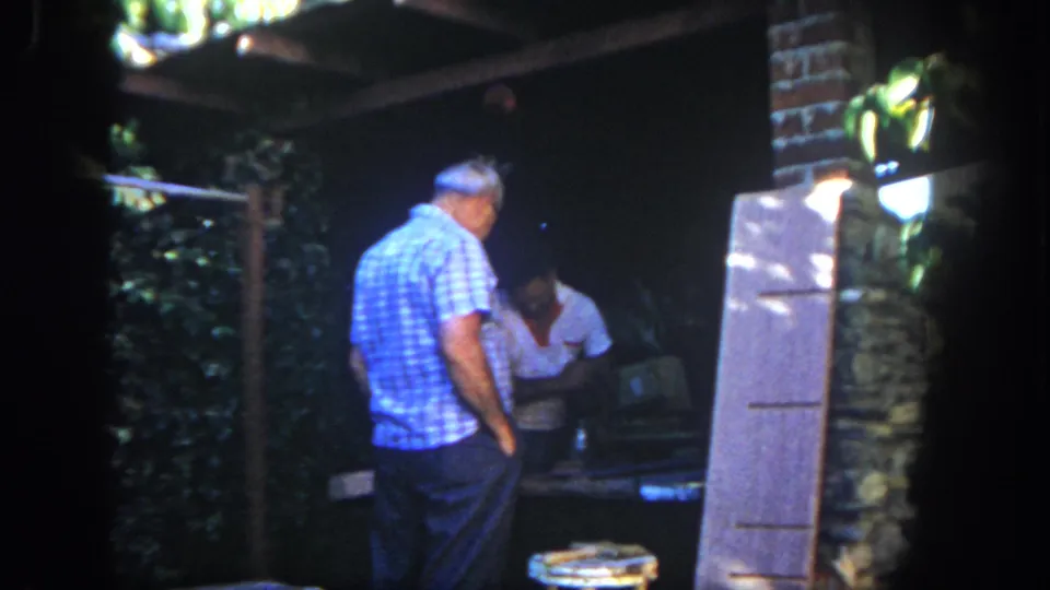1962 San Diego home-movie frame of two adult men in a shaded outdoor workshop with stools, boards, foliage, and a stone or brick wall.