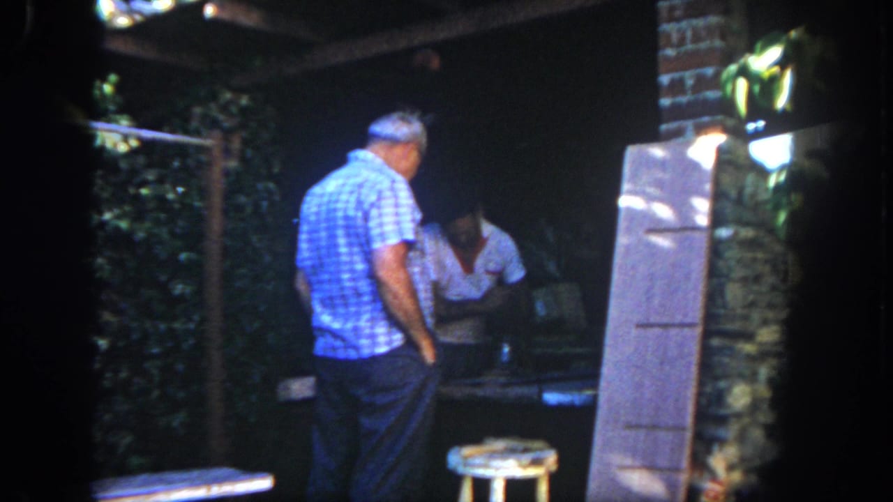 In a 1962 San Diego home-movie frame, a man in a blue checkered shirt stands on a patio near another adult and a grill area.