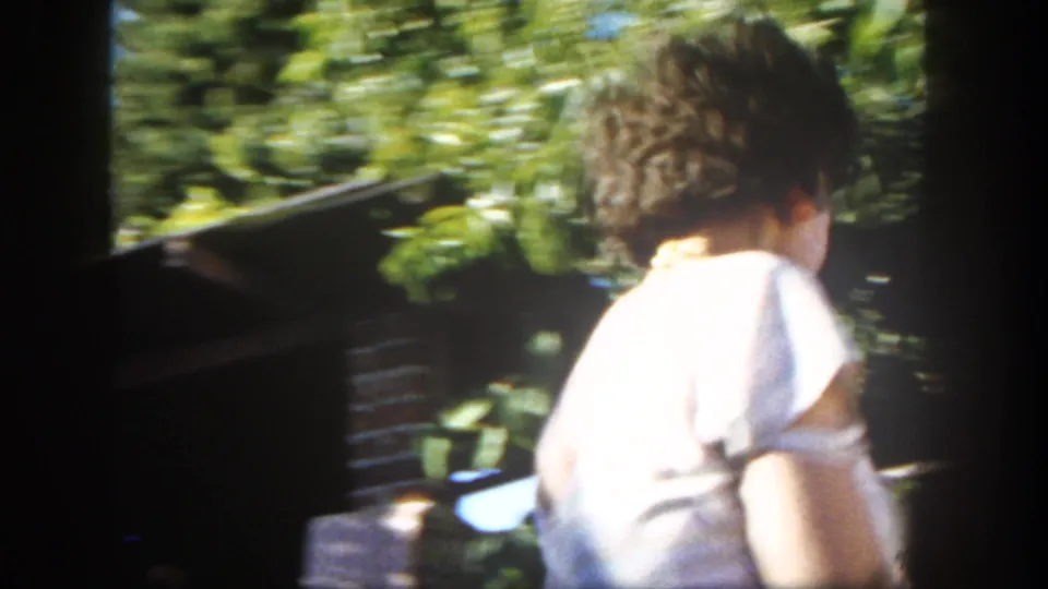 The handheld 8mm frame catches a woman in white from behind as she moves across the backyard patio near greenery.