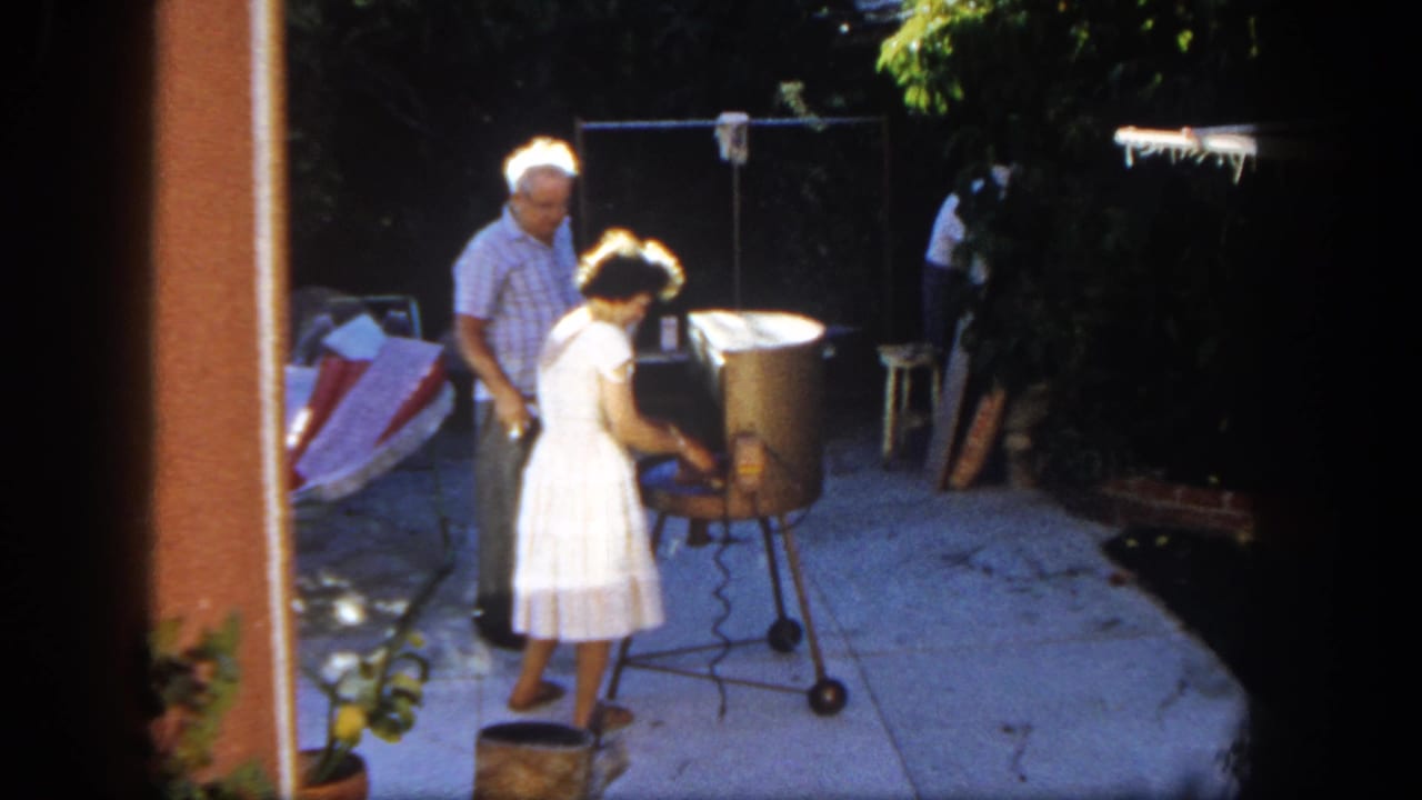 Woman in a white dress tends a round grill while a man stands beside her on a backyard patio.