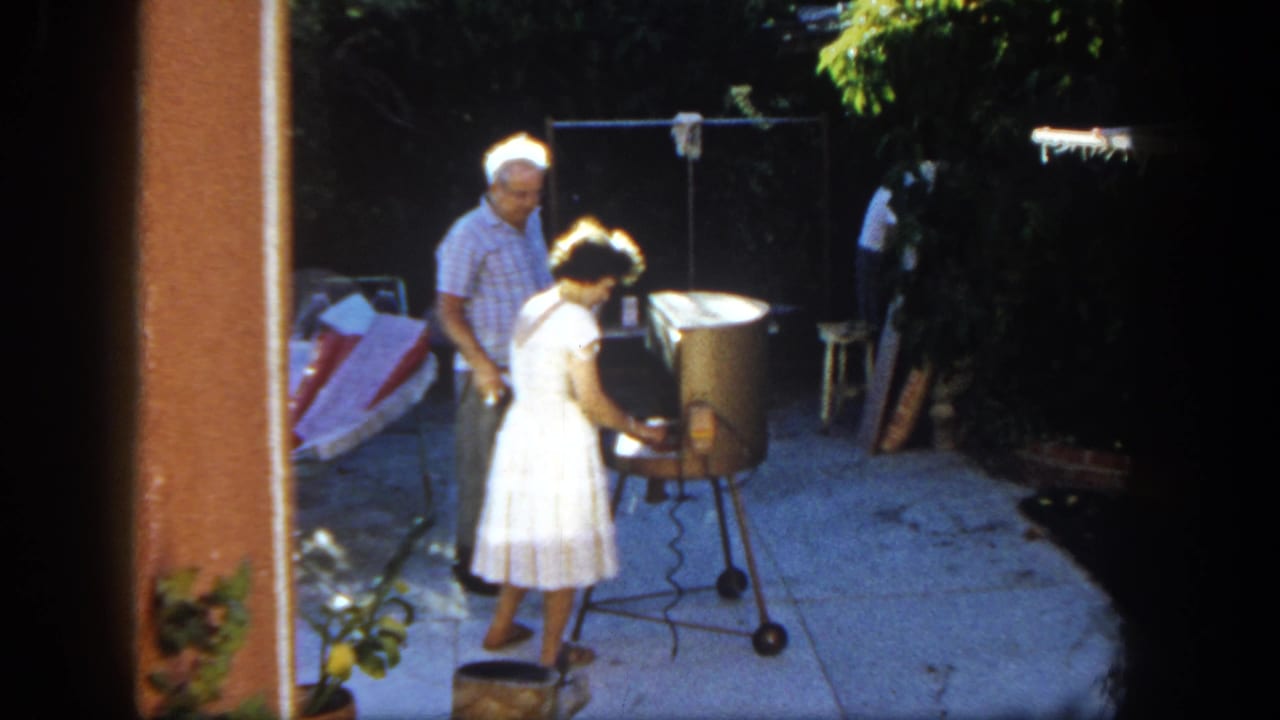 The woman continues tending the grill on a San Diego patio, with the man standing slightly behind her.