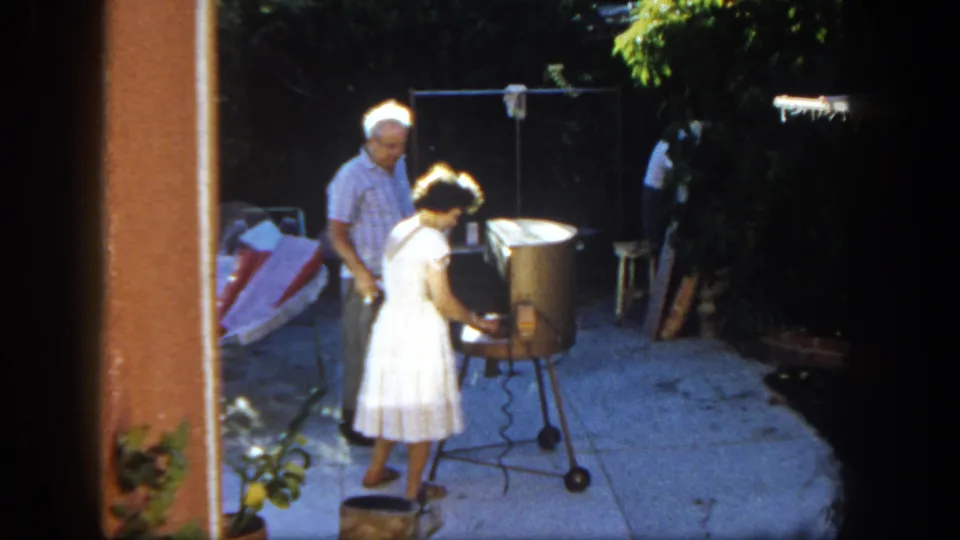 The woman continues tending the grill on a San Diego patio, with the man standing slightly behind her.