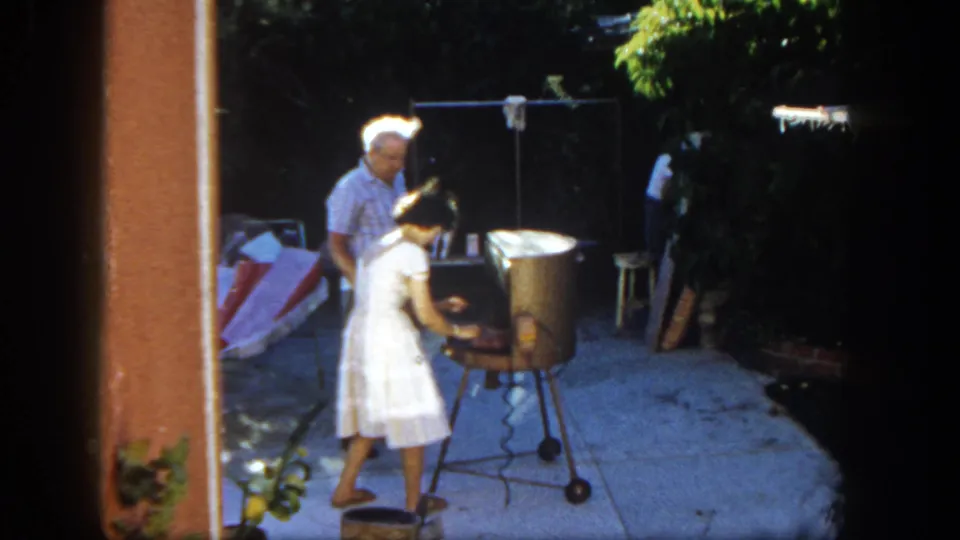 A 1962 backyard cookout moment shows the woman leaning near the grill while the man remains beside her.