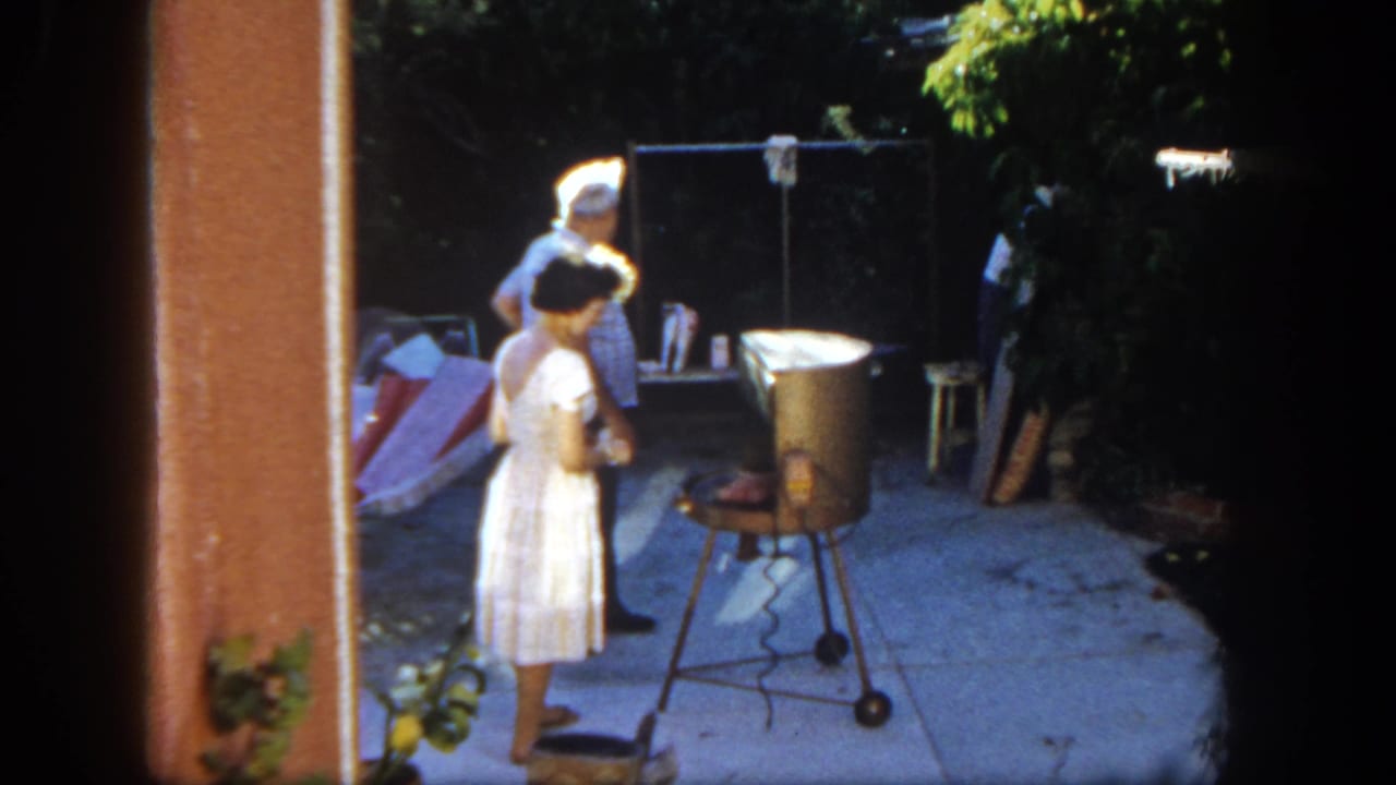 1962 San Diego home-movie frame showing a woman near a portable grill with a man nearby on a concrete backyard patio.