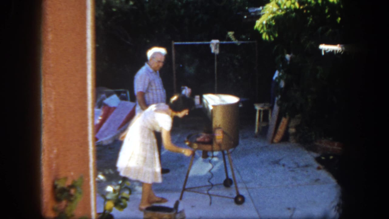 1962 San Diego backyard frame with a woman tending a portable grill and a man standing nearby amid patio furniture and greenery.
