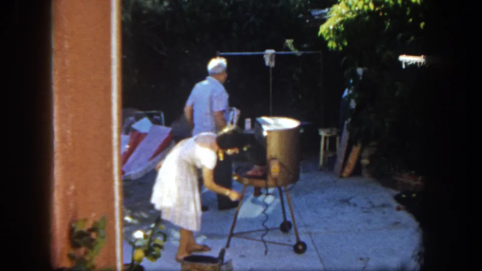 Home-movie frame from 1962 San Diego showing a woman adjusting a backyard grill while a man moves away in the patio background.