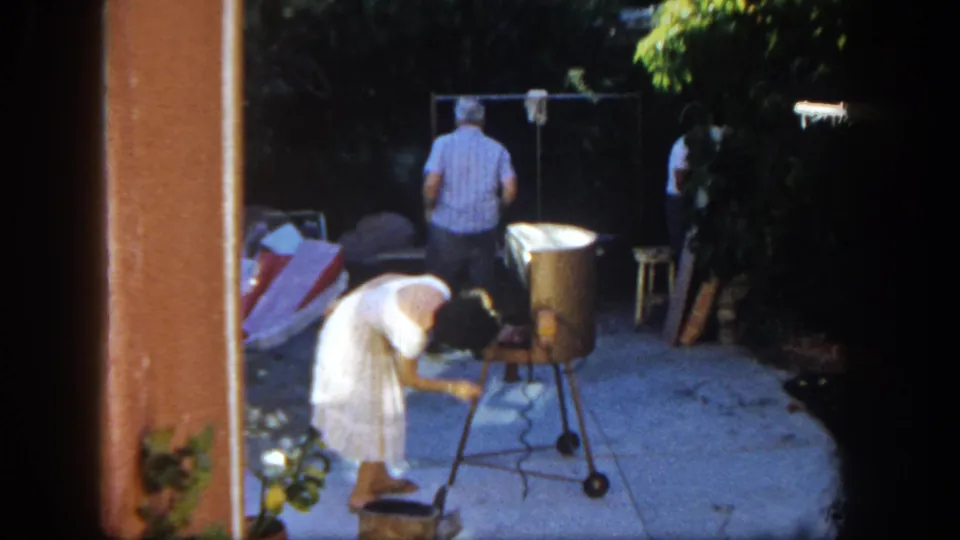 1962 San Diego backyard barbecue frame with a woman working at the grill and a man turned away beside a clothesline and patio items.