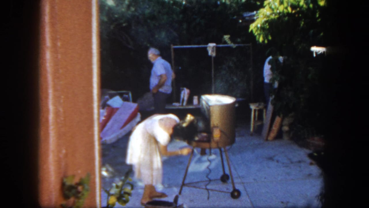 In 1962 San Diego home-movie footage, a young girl in a white dress tends a smoking metal grill on a backyard patio as an adult stands nearby.