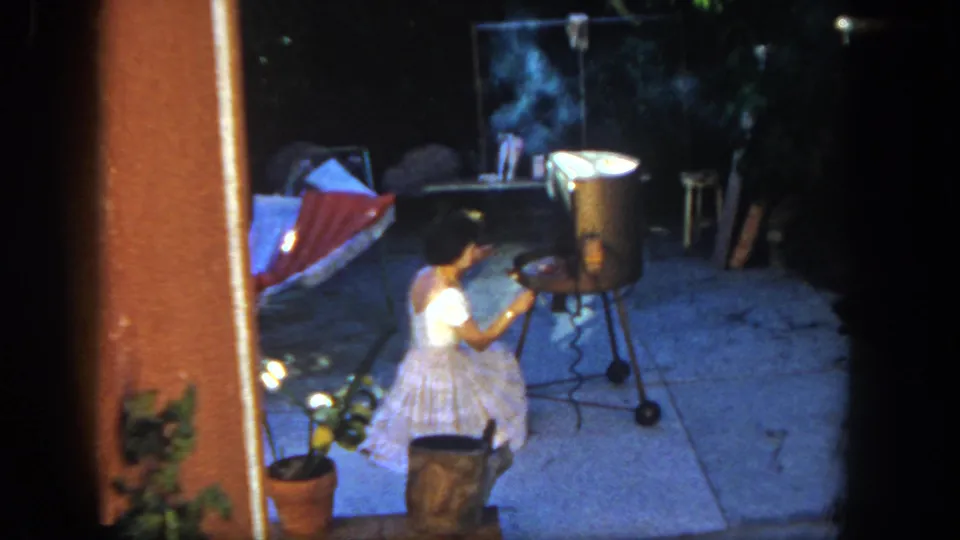 A young girl in a white dress remains beside the smoking backyard grill in this 1962 San Diego home-movie frame.