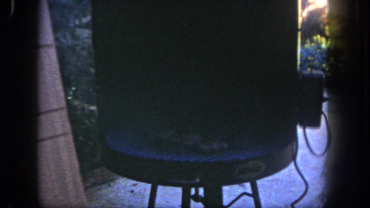 1962 San Diego home-movie frame showing a dark backyard view with a cylindrical grill, leafy plants, and strong 8mm frame shadows.