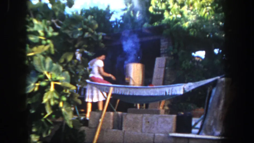 1962 San Diego 8mm frame showing a person in a light dress and red fabric tending a smoking backyard grill behind a hammock.