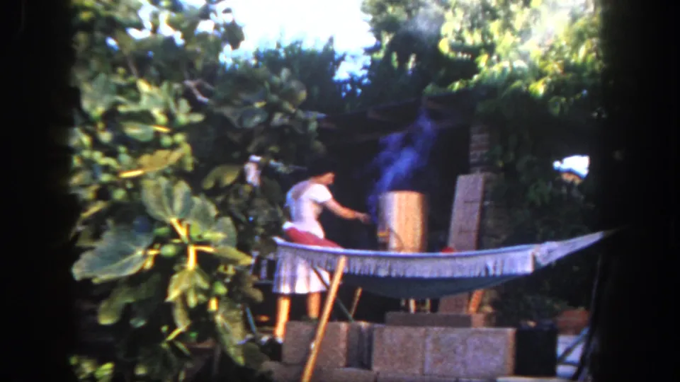 1962 San Diego home-movie frame with a person reaching toward a smoking cylindrical grill in a lush backyard with a hammock.