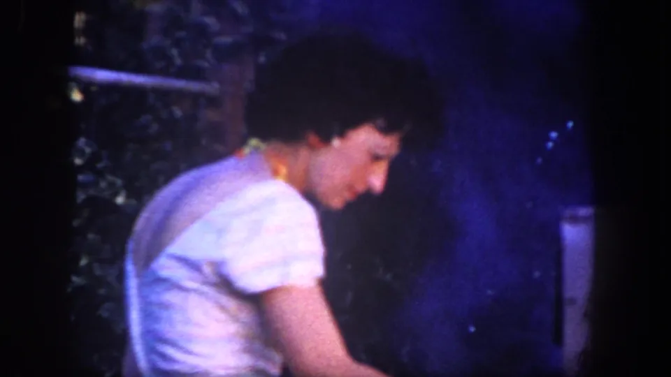 Close 1962 home-movie view of the woman in a white dress leaning toward smoke in the San Diego garden cooking scene.