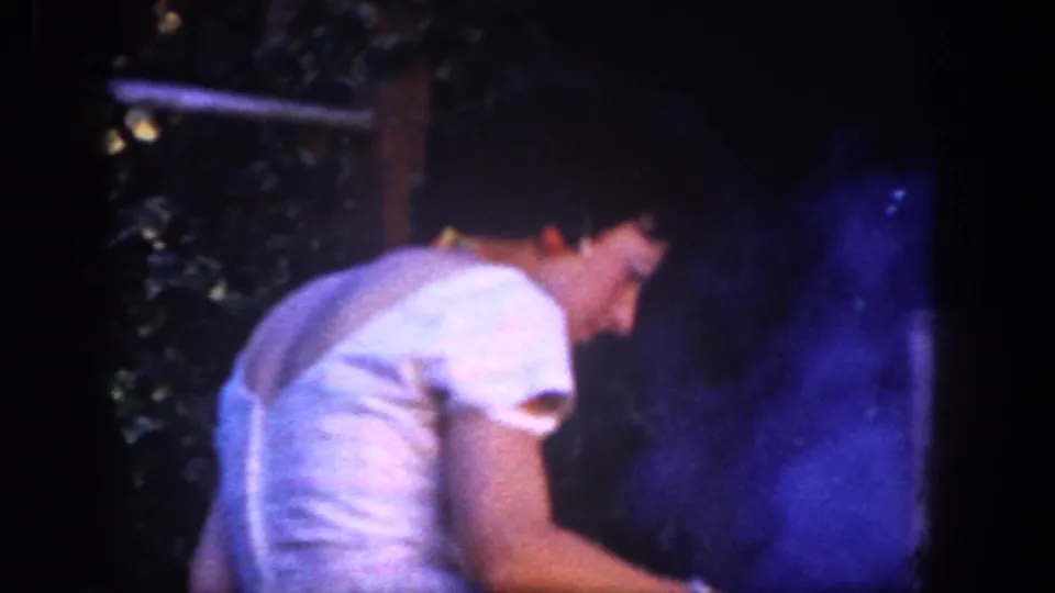 Close San Diego 1962 frame showing the woman turned toward the smoky outdoor cooking setup amid dense garden foliage.