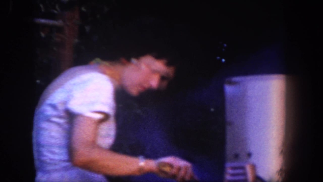 Woman in a white dress bends over an outdoor barbecue in dim nighttime light.