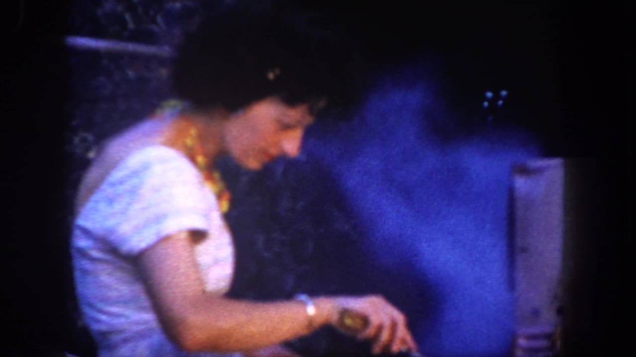 A woman in a white dress and gold necklace cooks beside blue smoke rising from a nighttime barbecue in San Diego, 1962.