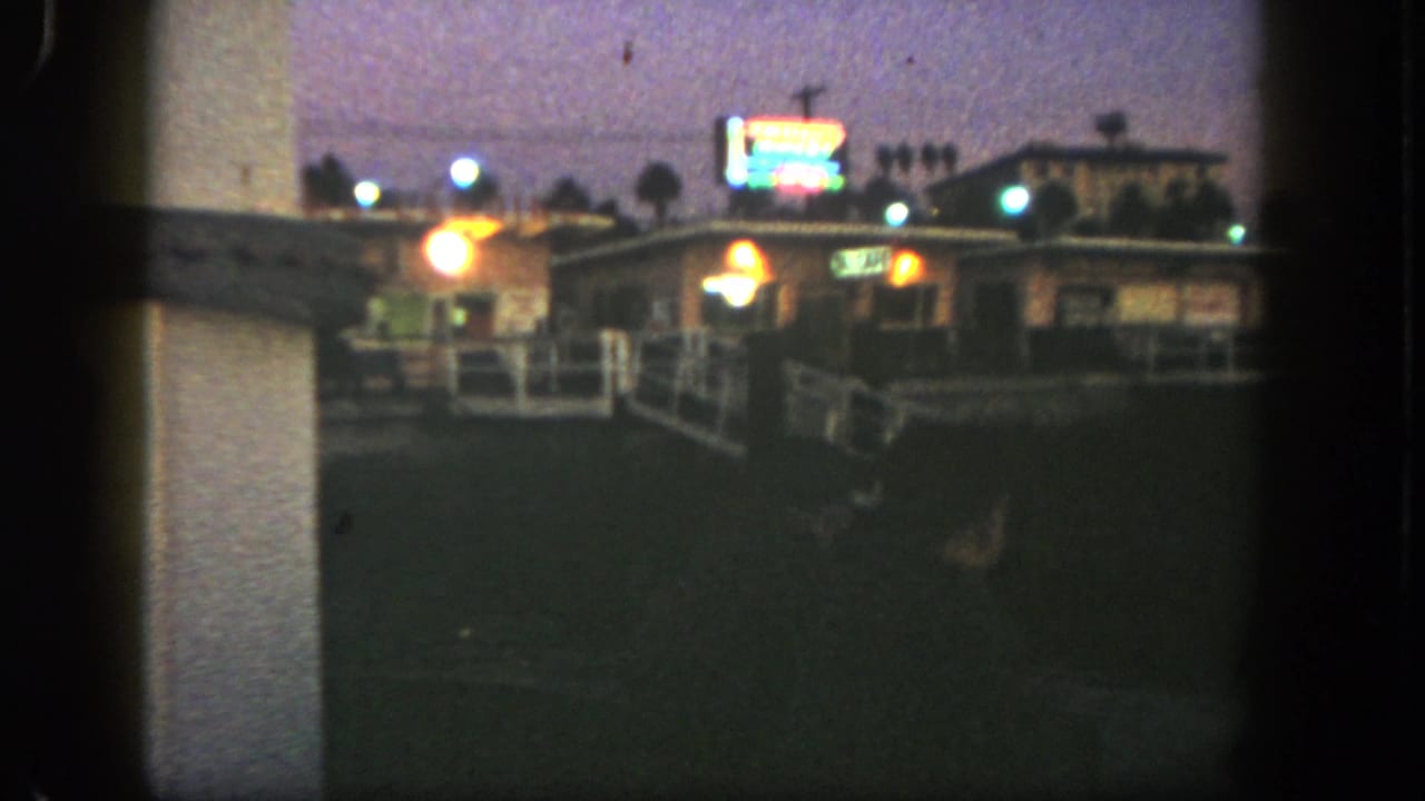 1962 San Diego home-movie frame showing a dim low building, neon glow, white railing, and palm silhouettes at dusk.