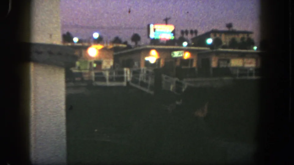 1962 San Diego home-movie frame showing a dim low building, neon glow, white railing, and palm silhouettes at dusk.