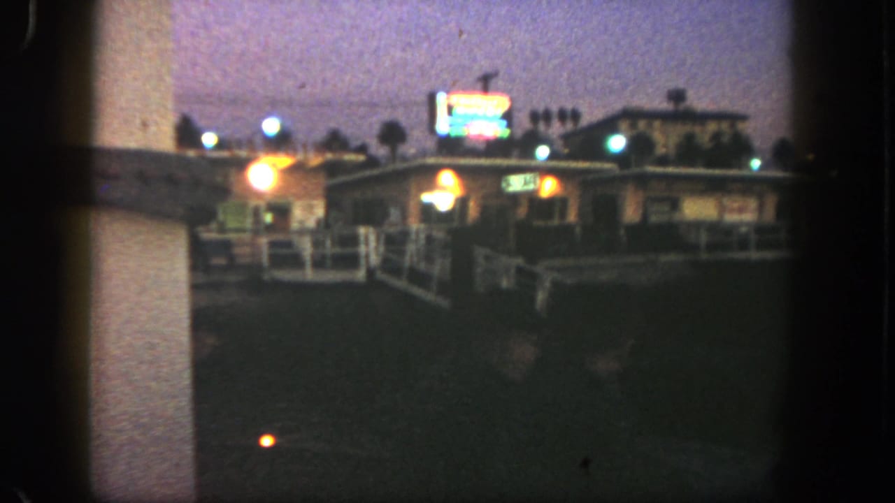 1962 San Diego frame with a blurred neon sign above a low roadside building, warm lights, railings, and palms.