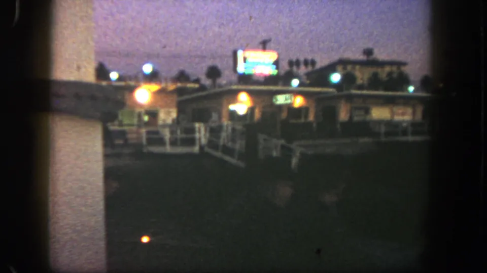 1962 San Diego frame with a blurred neon sign above a low roadside building, warm lights, railings, and palms.