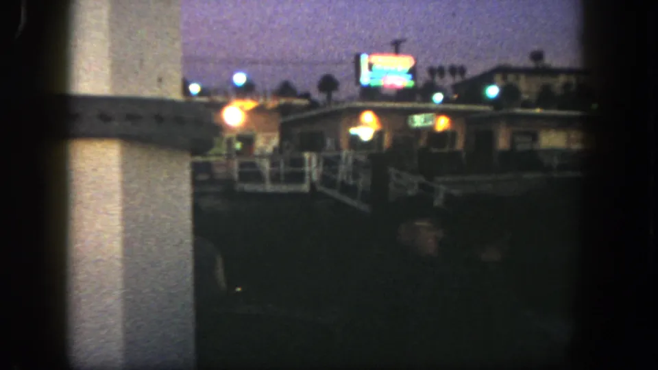 1962 San Diego frame of a dim exterior scene with a low building, bright sign, white railing, and palms.