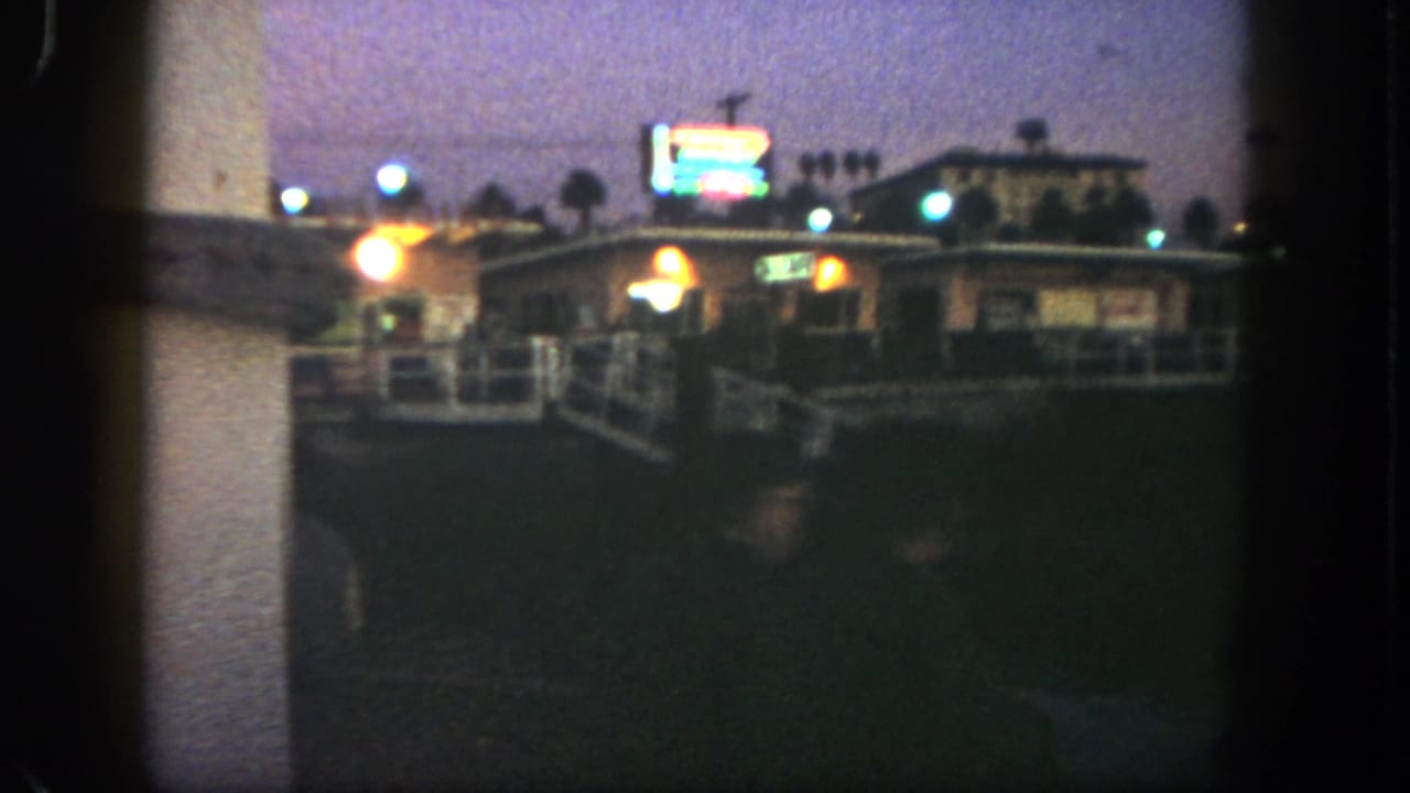 1962 San Diego home-movie frame showing a dim commercial exterior, neon-like signage, streetlights and railing at dusk.