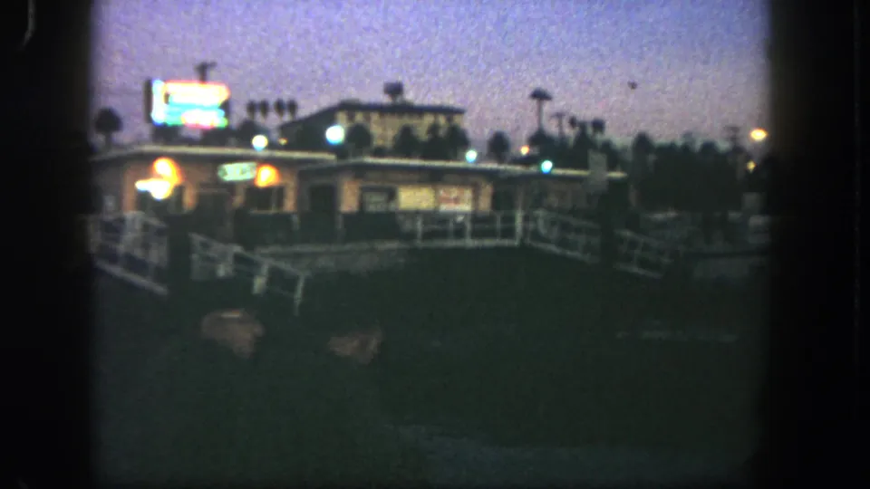 1962 San Diego 8mm frame showing a low commercial structure with glowing signs, white railing and dark foreground at evening.