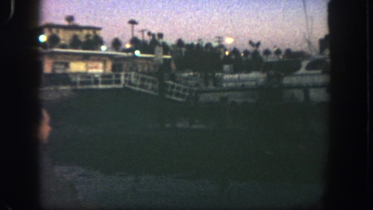 Dusk marina view with a docked white yacht, pier railings, lamps, palm trees, and a small waterfront building.
