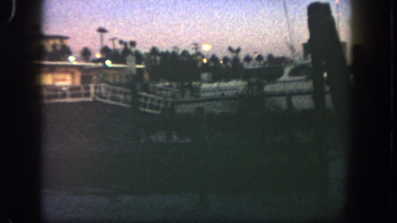 1962 San Diego home-movie frame with a white yacht docked near railings, warm harbor lights, palm trees, and dark water under a fading sky.