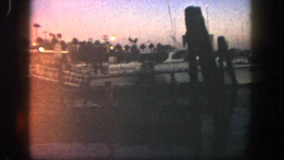 1962 San Diego 8mm frame showing a yacht at a pier, soft orange dusk light, palm silhouettes, water reflections, and possible distant people or posts.