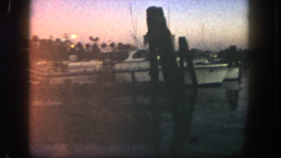 1962 San Diego home-movie frame of a docked yacht at twilight, with railings, warm lights, palms, water reflections, and dark silhouetted shapes at right.