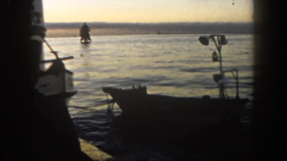 1962 San Diego harbor scene with dark dock posts, a small boat in the foreground, calm water, and a distant vessel on the horizon.