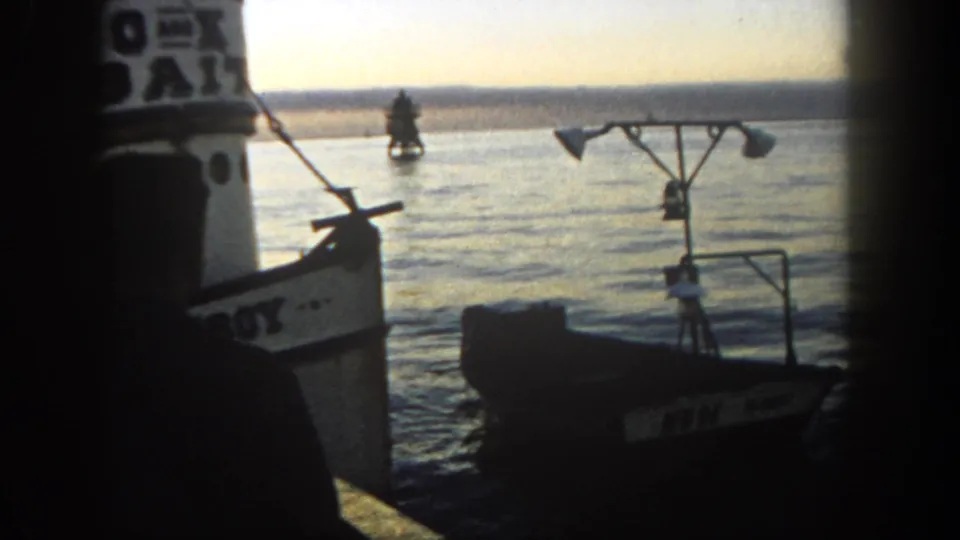 A small empty boat drifts beside a larger vessel in 1962 San Diego harbor, with a buoy visible across the water.