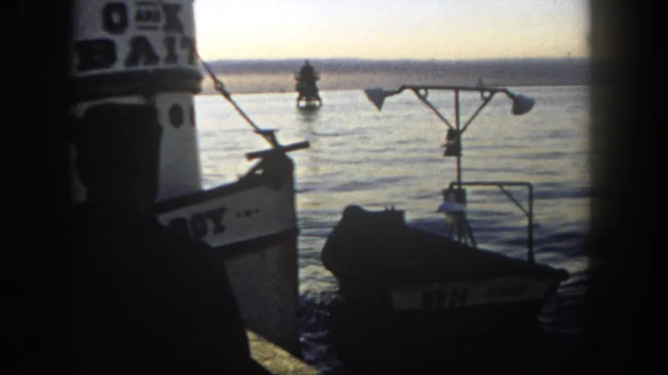 A silhouetted young sailor watches a small empty boat in San Diego harbor, filmed on 8mm in 1962.