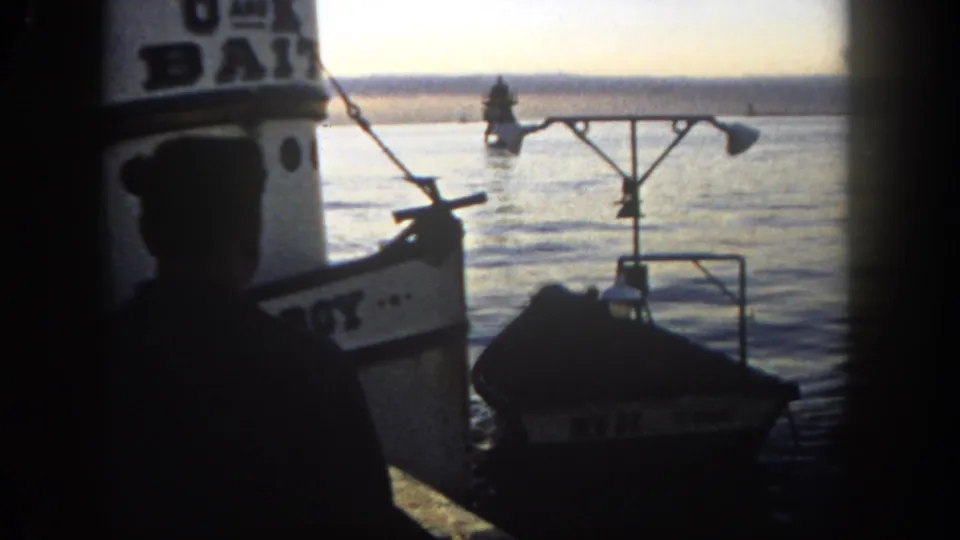 1962 San Diego harbor frame with a silhouetted sailor, small empty boat, docked vessel, and buoy across the water.