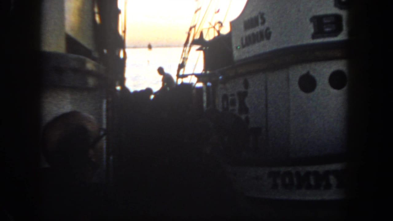 1962 San Diego harbor footage showing two close boats, dark dockside figures, rigging, and sunset glare on the water.