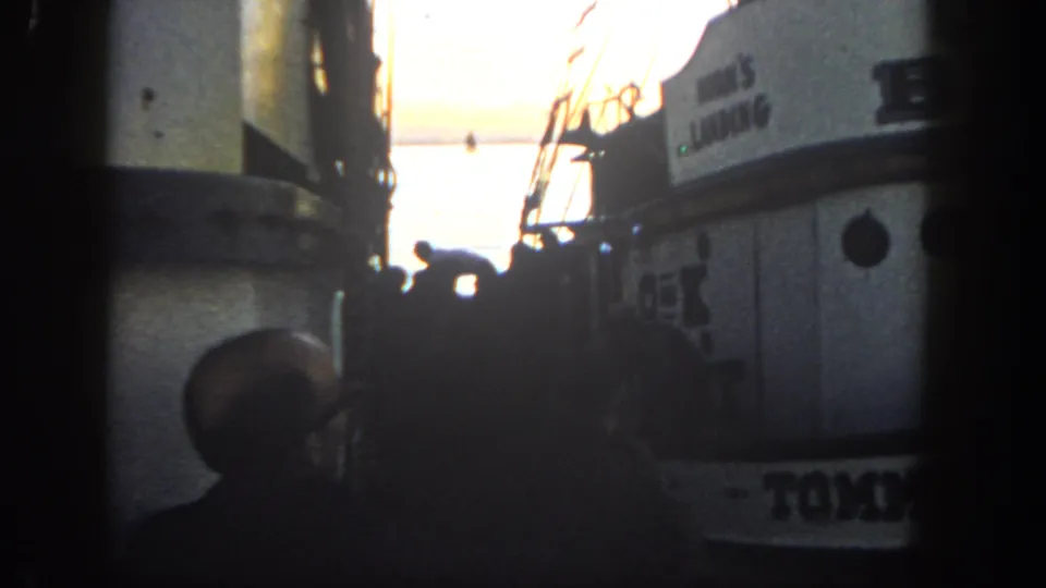 1962 8mm harbor frame with a silhouetted person near the dock, close boat hulls, rigging, and the visible name TOMMY.