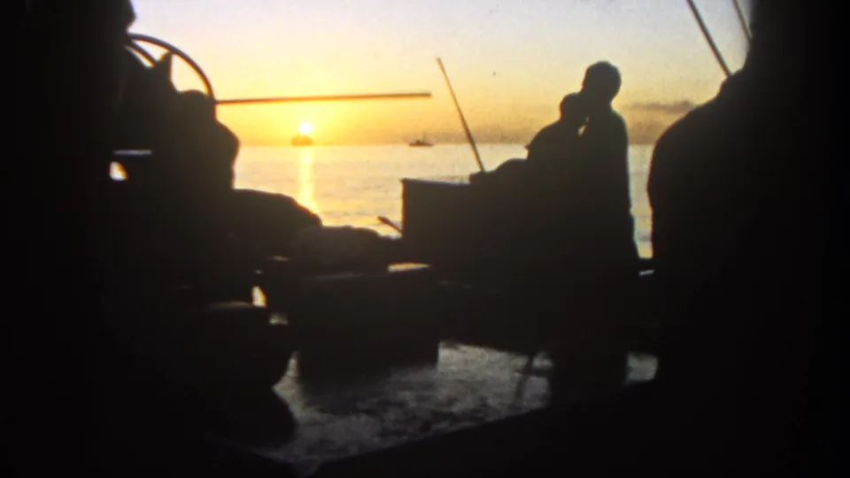 1962 San Diego-area home-movie frame with two dark figures near rods and deck equipment, filmed into the sunset over open water.
