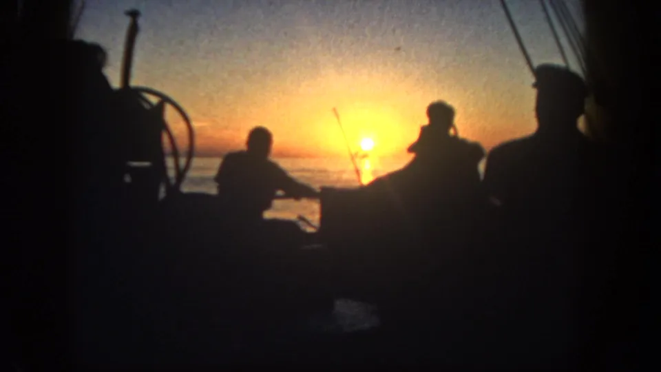 Home-movie frame of silhouetted adults working around boat gear as the low sun glows over San Diego water in 1962.