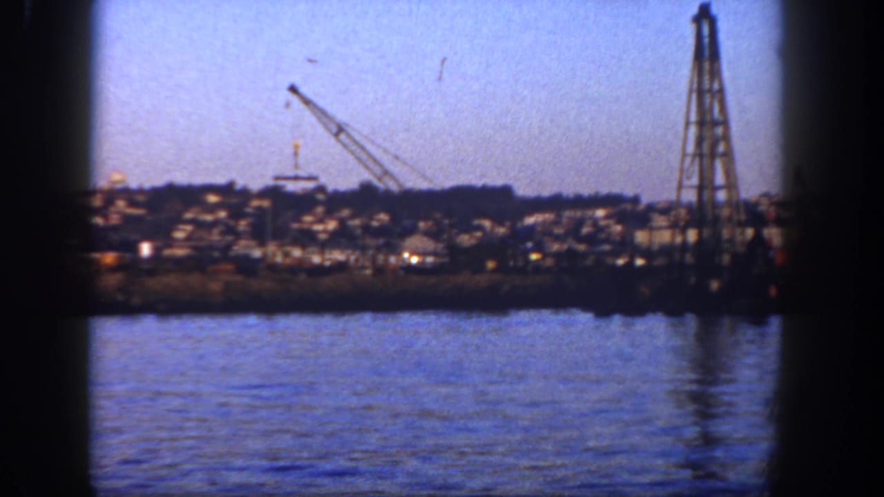 1962 San Diego 8mm view across water toward a shoreline crane, tall industrial tower, and houses on a low hillside.