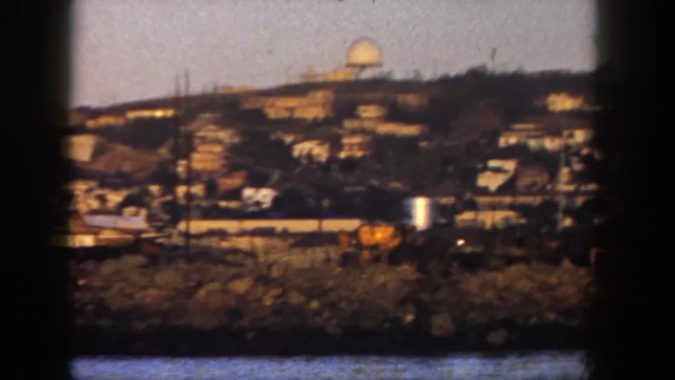 Closer 1962 San Diego 8mm view of hillside houses above shoreline structures, with utility poles and a rounded light-colored ridge feature.