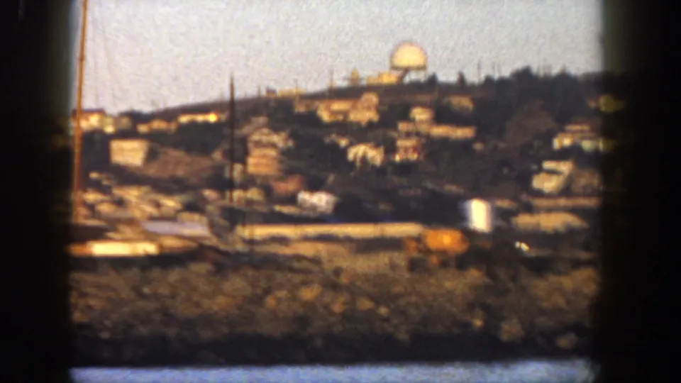 1962 San Diego home-movie frame showing hillside homes above a shoreline area, with utility poles and dry foreground vegetation.