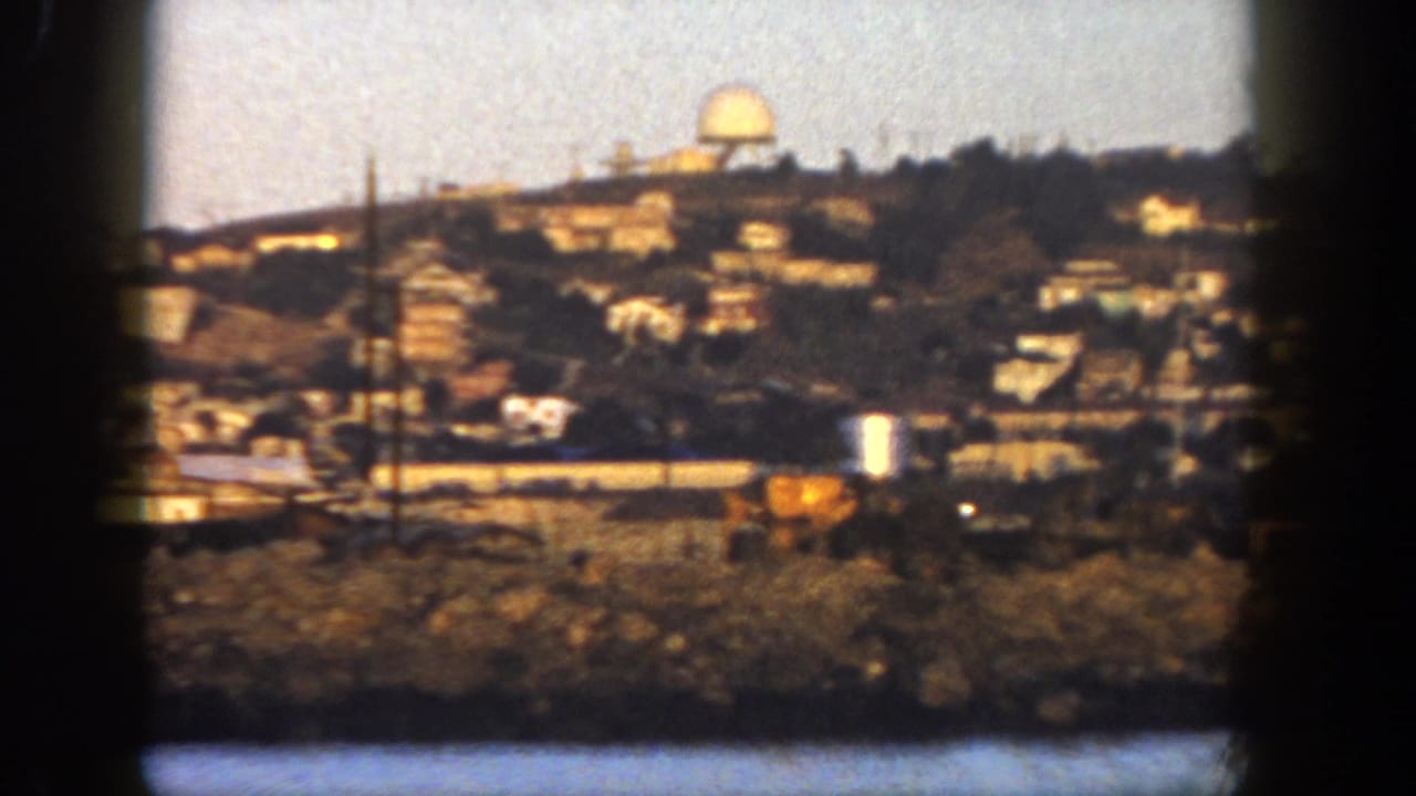 1962 San Diego coastal view with hillside buildings, utility poles, rocky waterfront, and a distant dome-like structure.