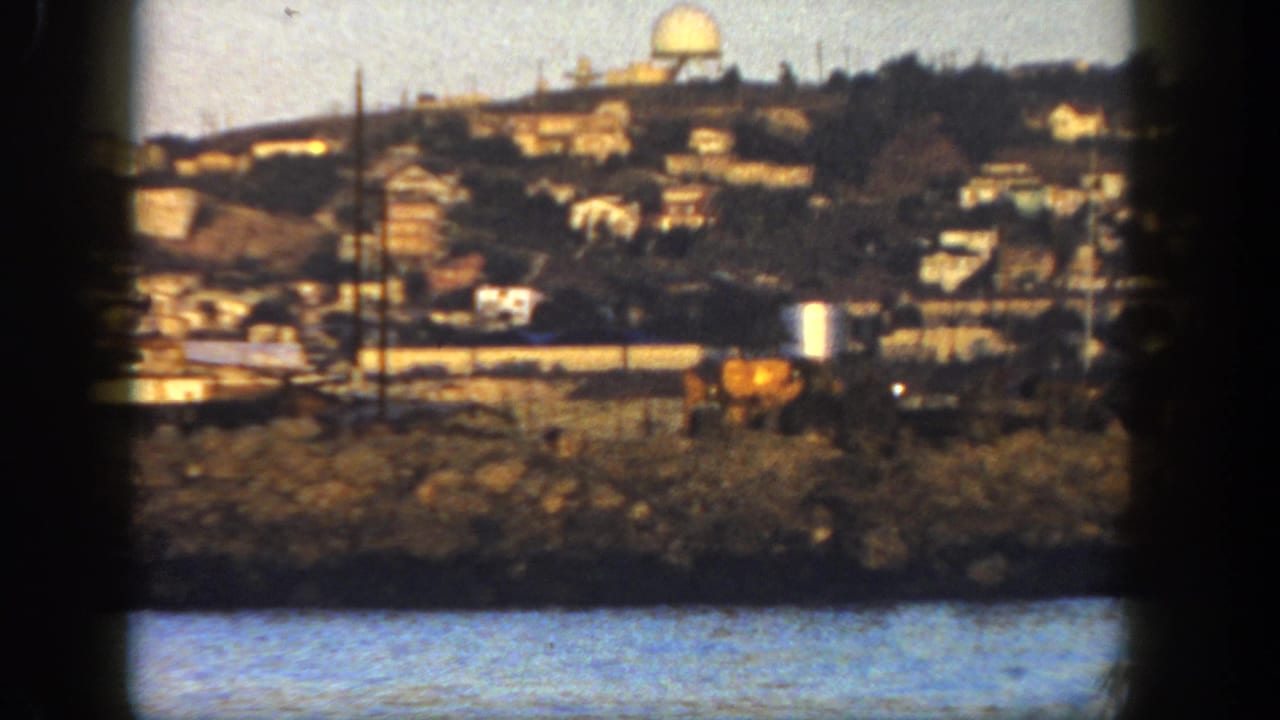 1962 San Diego shoreline view showing hillside homes, a distant dome-like structure, rocky waterfront, and a yellow vehicle.