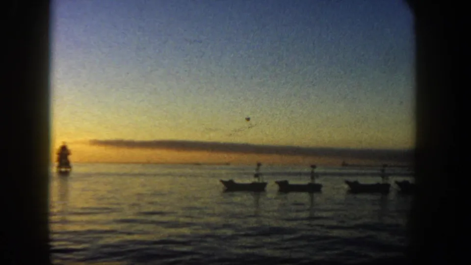 1962 San Diego 8mm frame showing small boats in silhouette on calm water at sunset, with a distant lighthouse or marker near the horizon.